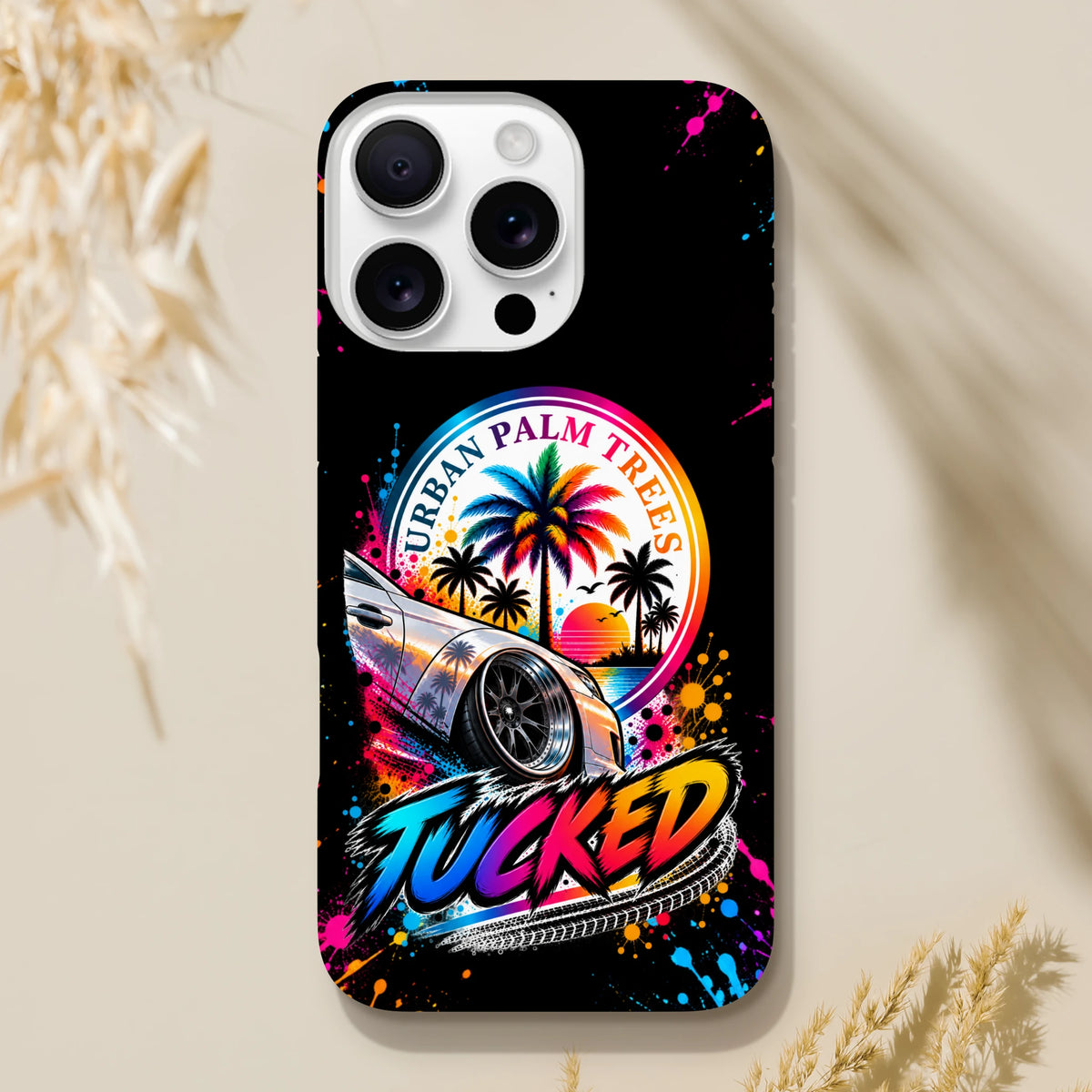 Tucked Petrolhead iPhone case featuring a lowered modified car, colorful Urban Palm Trees sunset graphic, and vibrant paint splatter art on black.