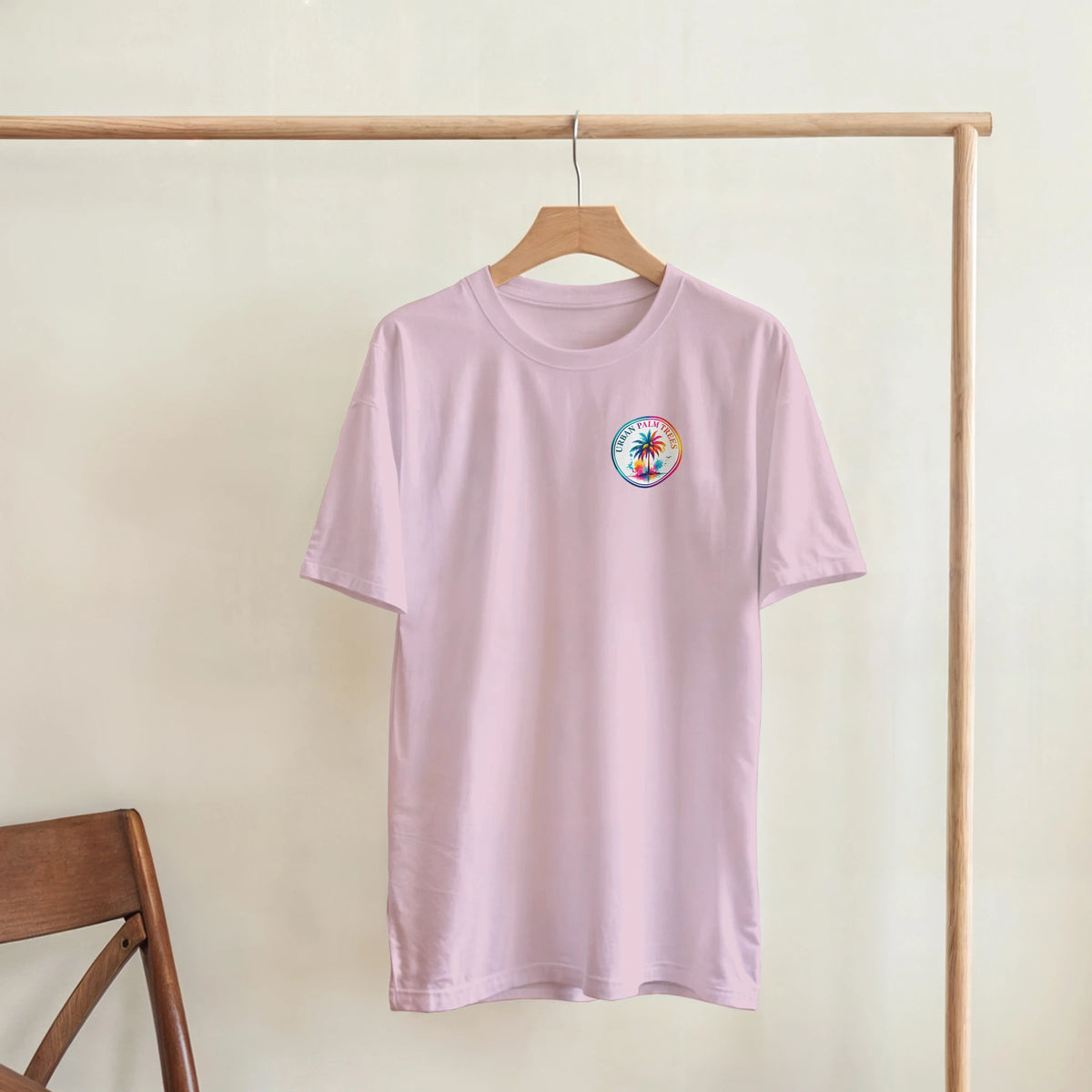 Pastel pink ladies tee with small colorful retro palm tree graphic on chest, hanging on wooden rack for casual style.