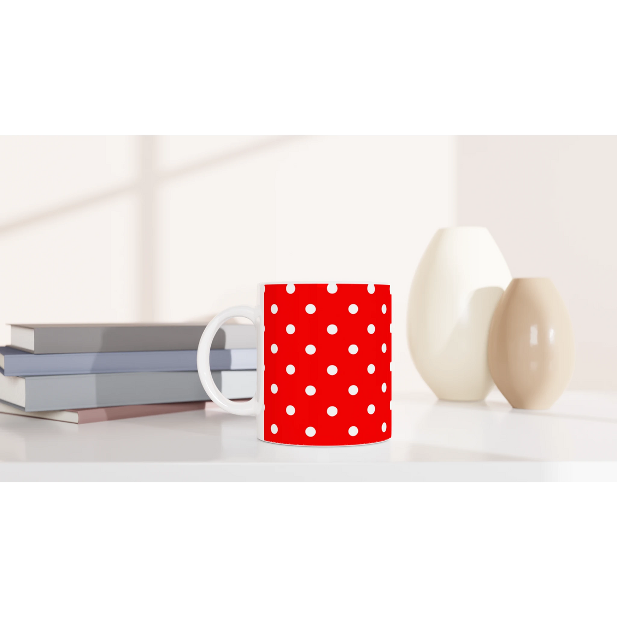 Red and white polka dot ceramic mug sitting on a white desk next to stacked books and decorative vases.