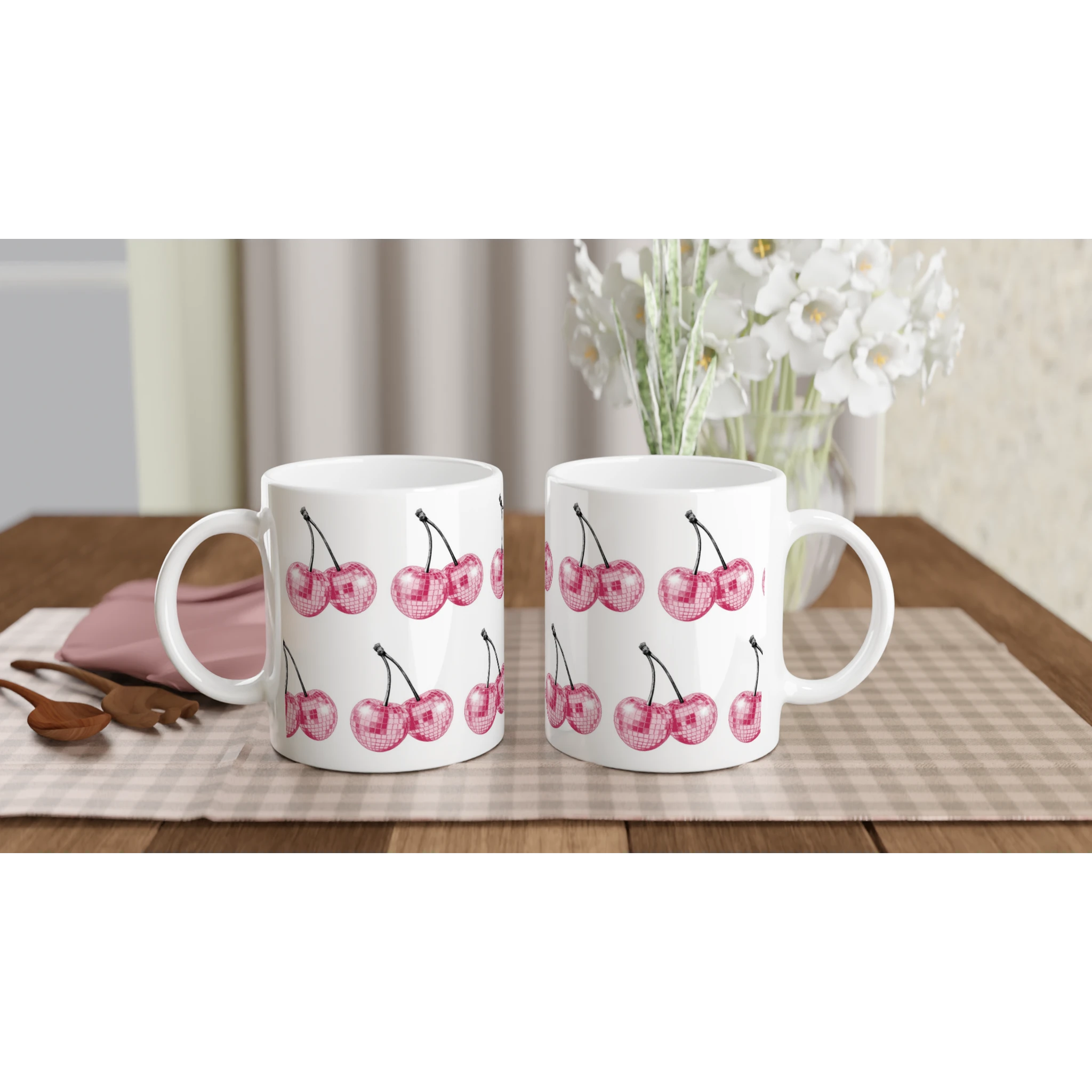 Two white ceramic Glitter Ball Cherries mugs on a checkered placemat with a vase of white flowers in the background.