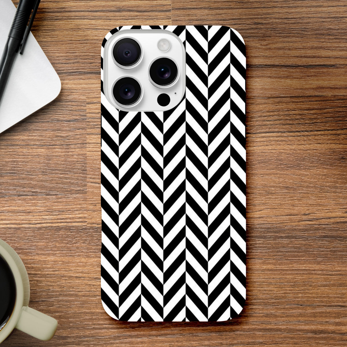 Black and white herringbone geometric iPhone case on wood desk. Tough dual-layer shock absorbing cover for iPhone 13-16 with glossy finish.