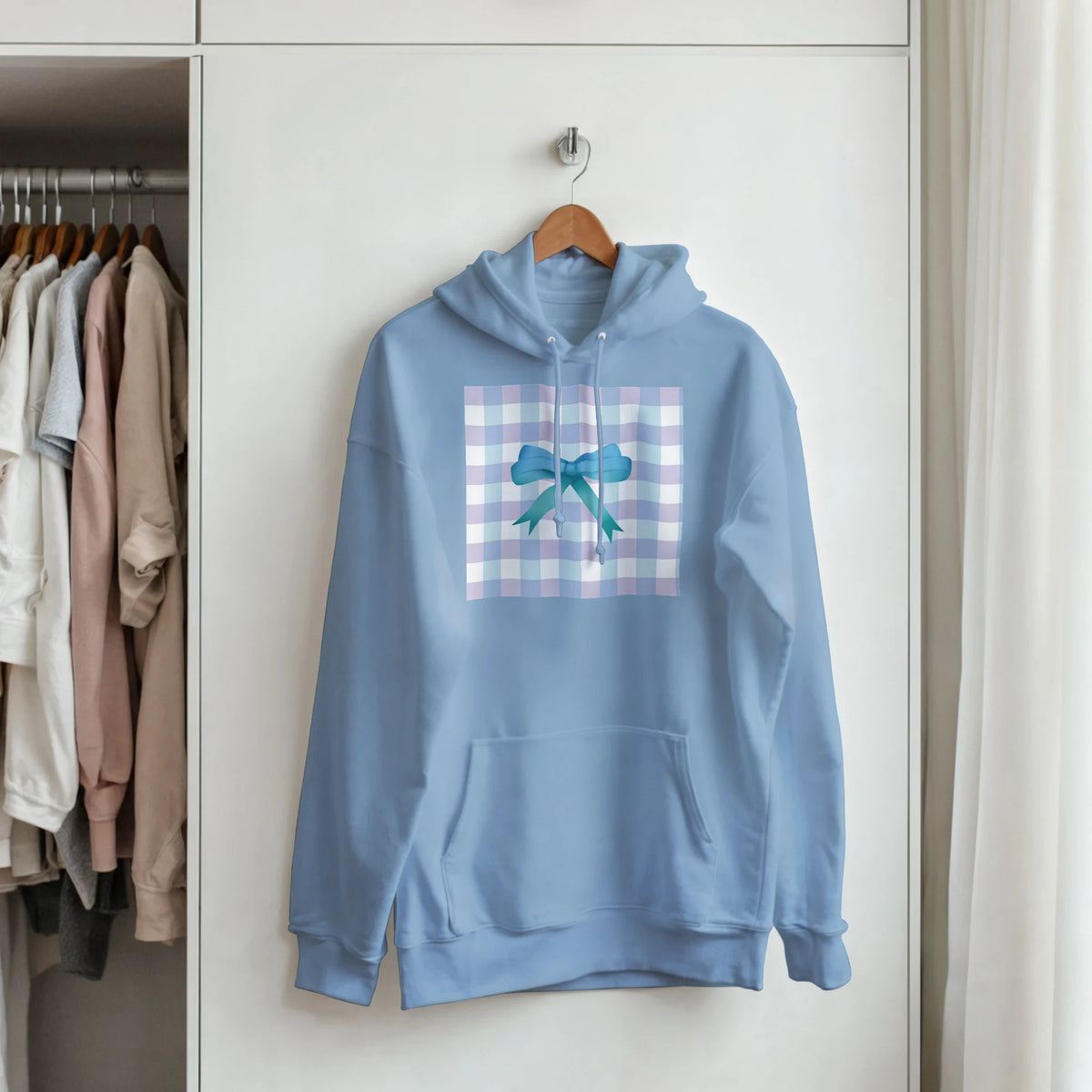Light blue coquette hoodie featuring a pastel gingham and blue bow graphic print, hanging on a wooden hanger in a closet.