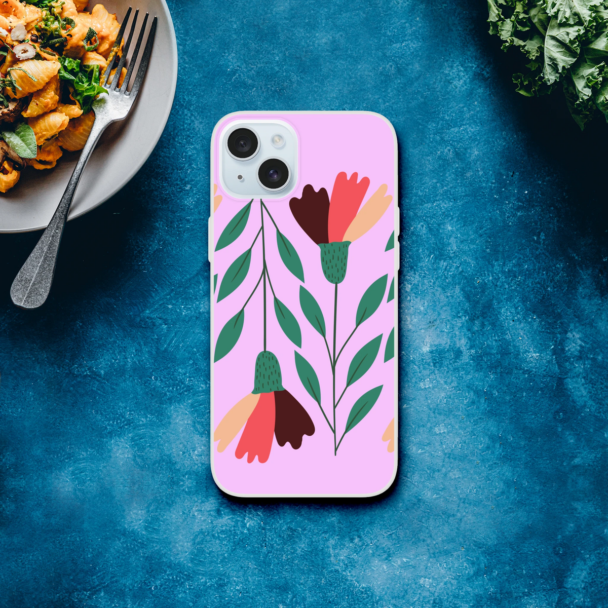 Pastel pink floral iPhone case with vintage botanical artwork and camera cutout on a blue surface beside a pasta plate