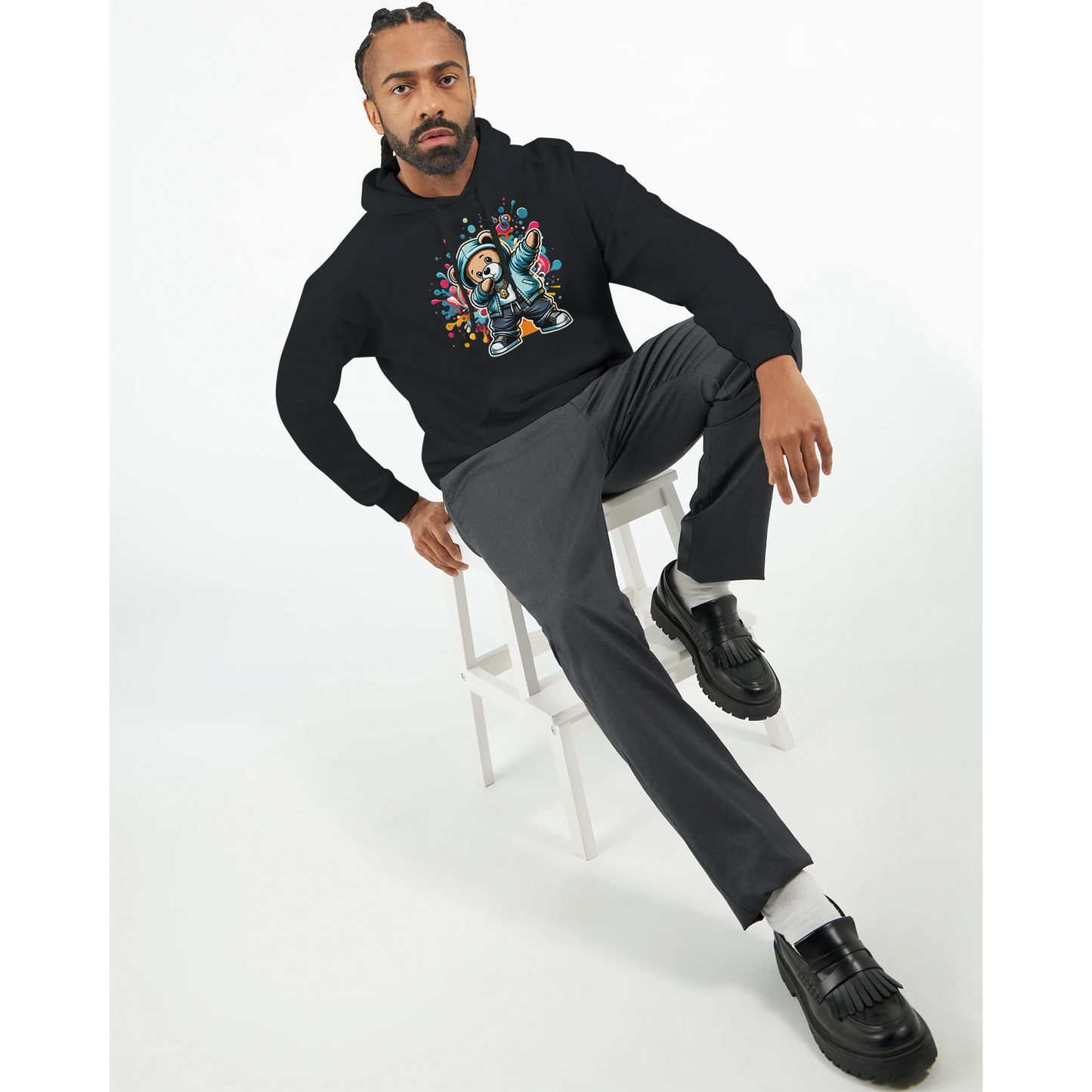 Man sitting on a stool wearing a black men's teddy bear hoodie with a colorful graphic design and gray pants. 5