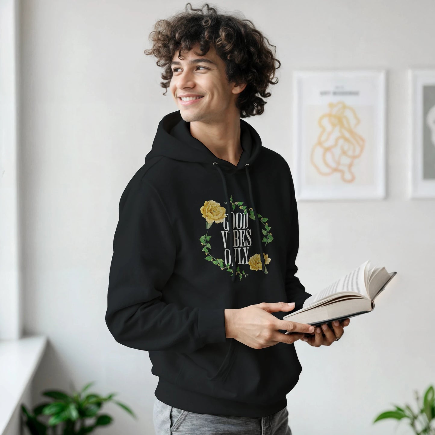 Man wearing black soft cotton blend Good Vibes Only graphic hoodie with floral wreath, holding an open book.