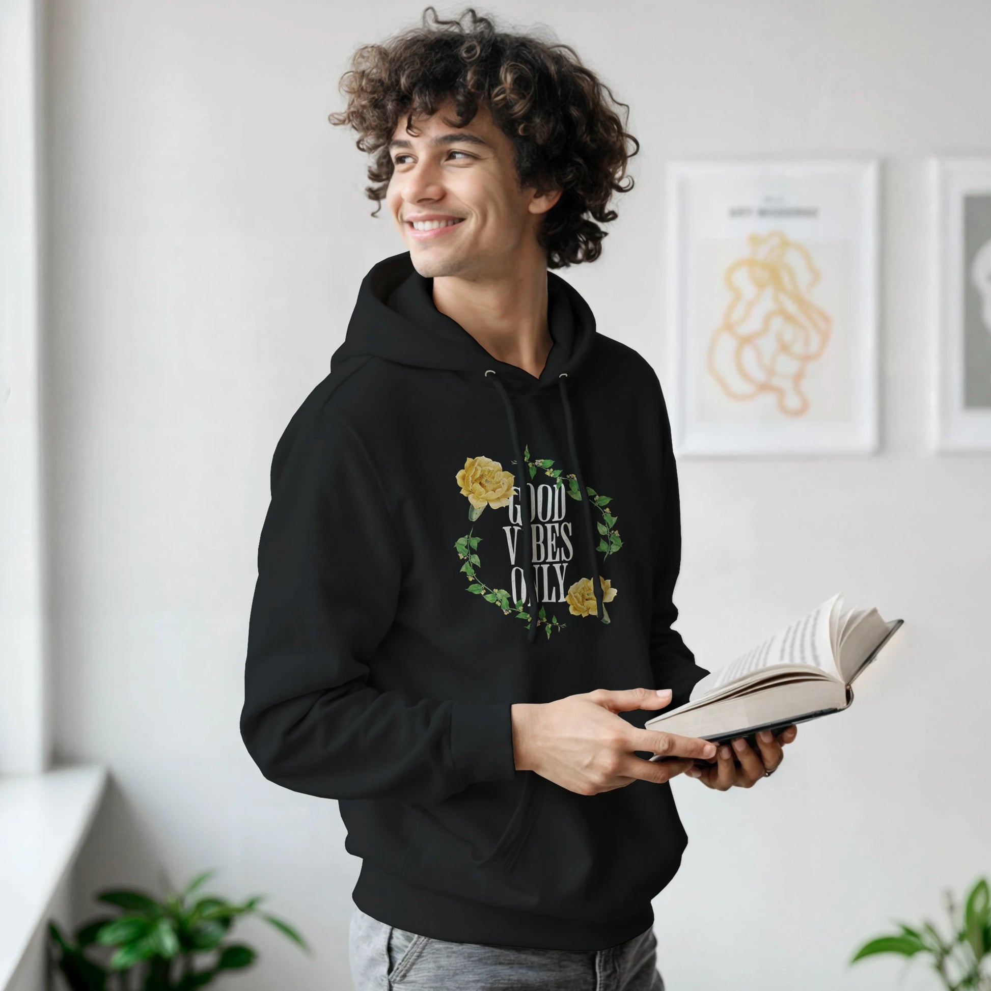 Man wearing black soft cotton blend Good Vibes Only graphic hoodie with floral wreath, holding an open book.