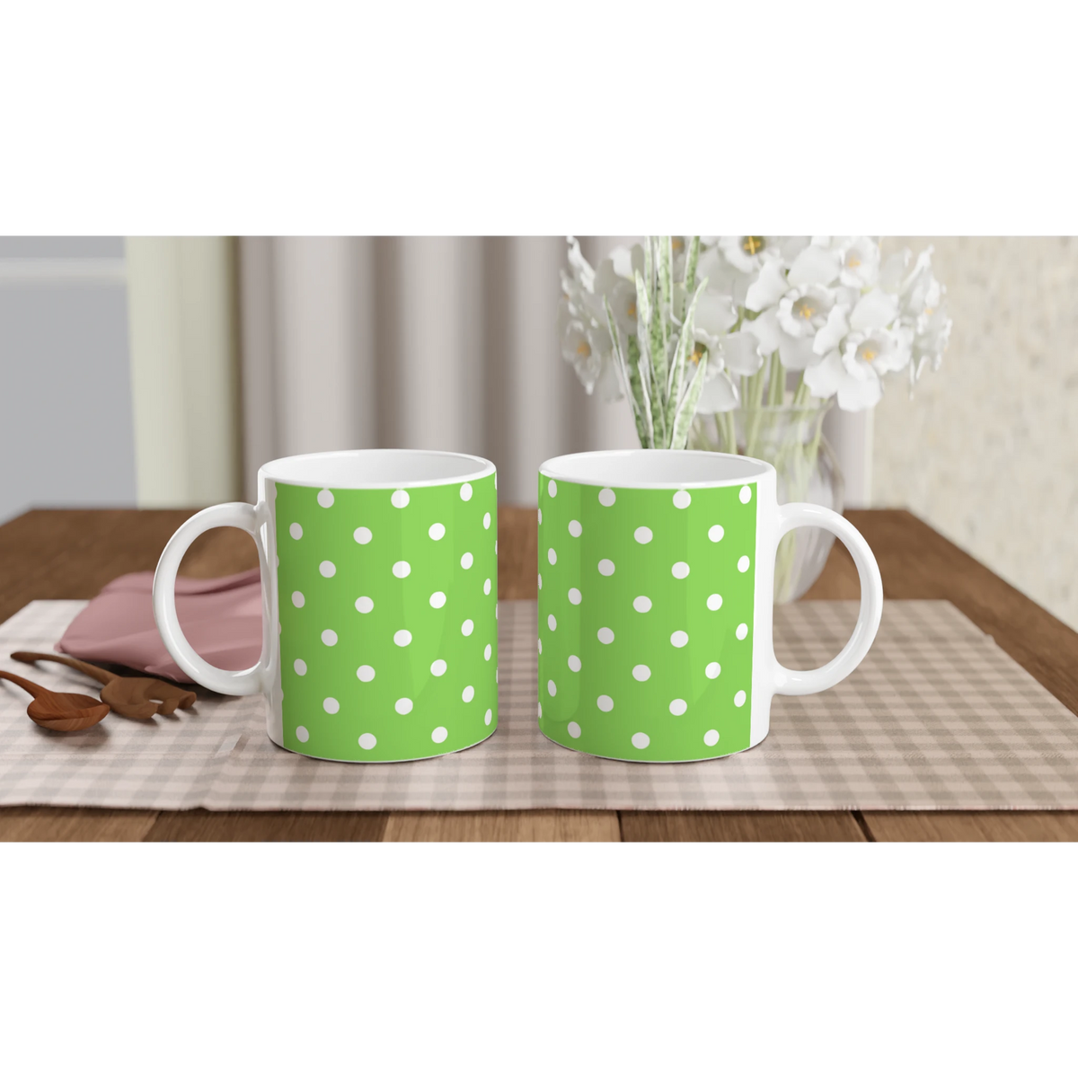 Two green ceramic mugs with white polka dots sit on a checkered placemat beside wooden spoons and white flowers.