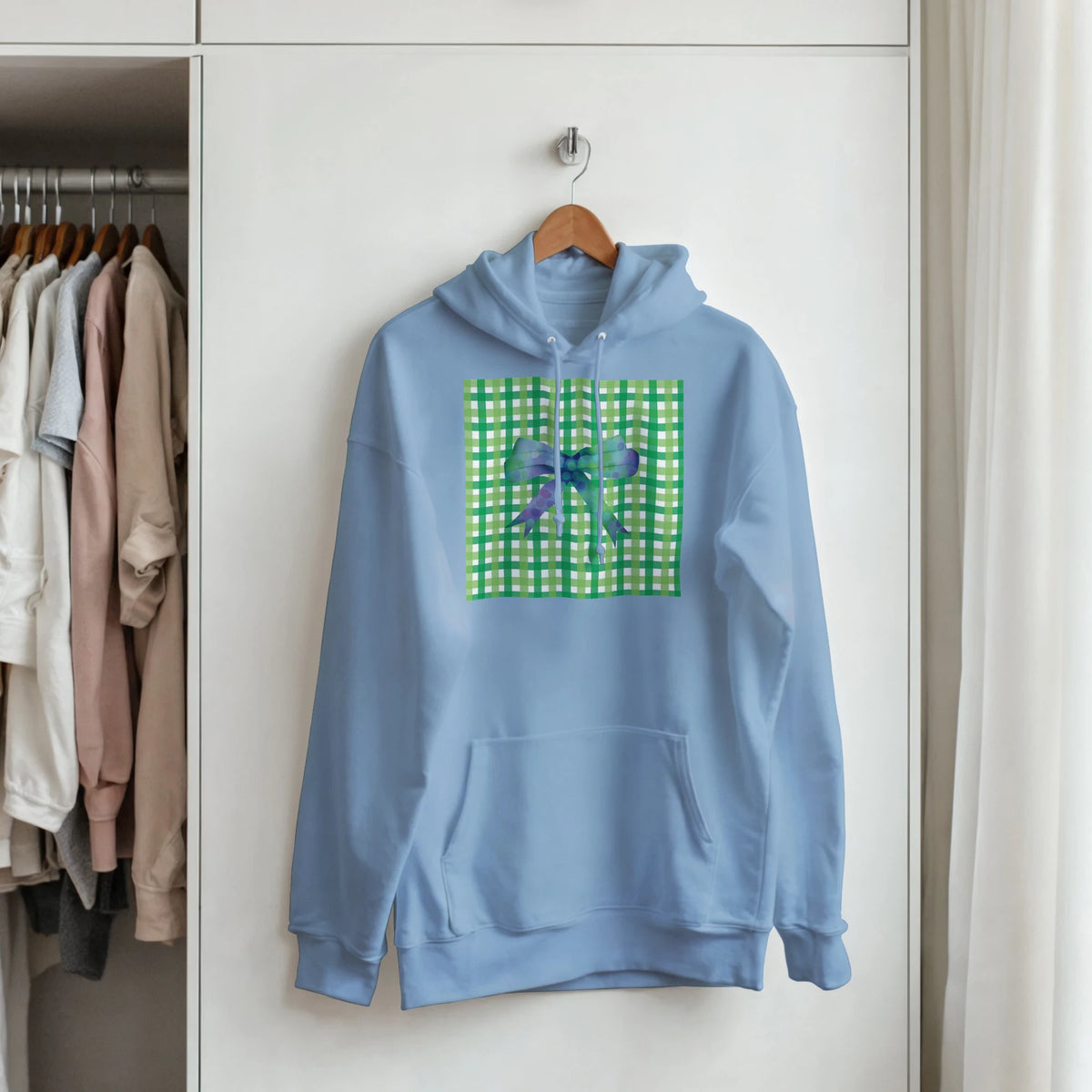 Blue pullover hoodie with a green gingham bow graphic on the chest, hanging on a hanger in a closet.