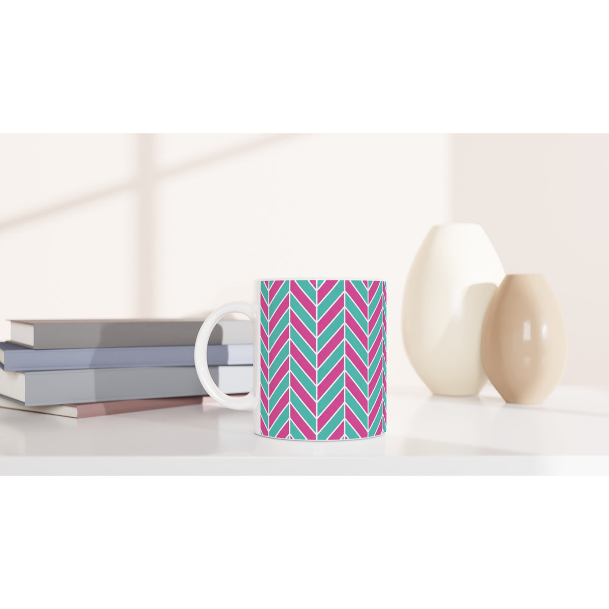 Pink and aqua herringbone patterned ceramic mug with glossy finish, sitting on a white desk beside stacked books and vases.