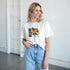 Smiling woman wears white Miami graphic T-shirt with tropical sunset and palm trees, paired with light-wash jeans.