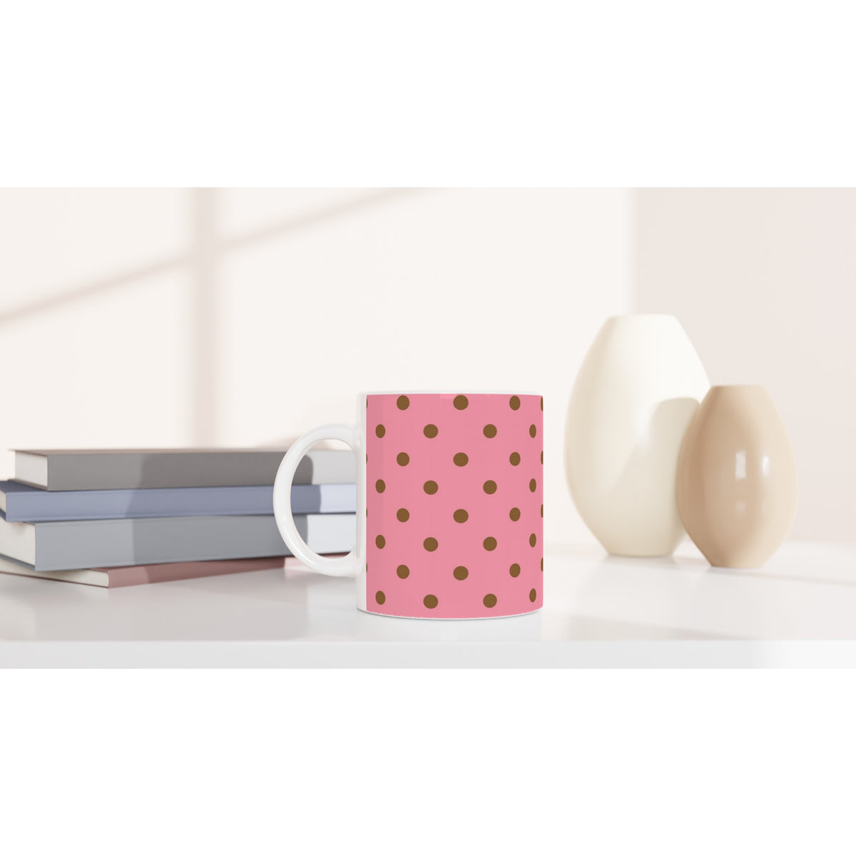 Pink and brown polka dot ceramic mug on a white desk beside books and vases. Retro spotty coffee cup for daily use.