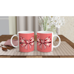Two pink mugs with a heart-shaped bow design on a table, white flowers in the background.