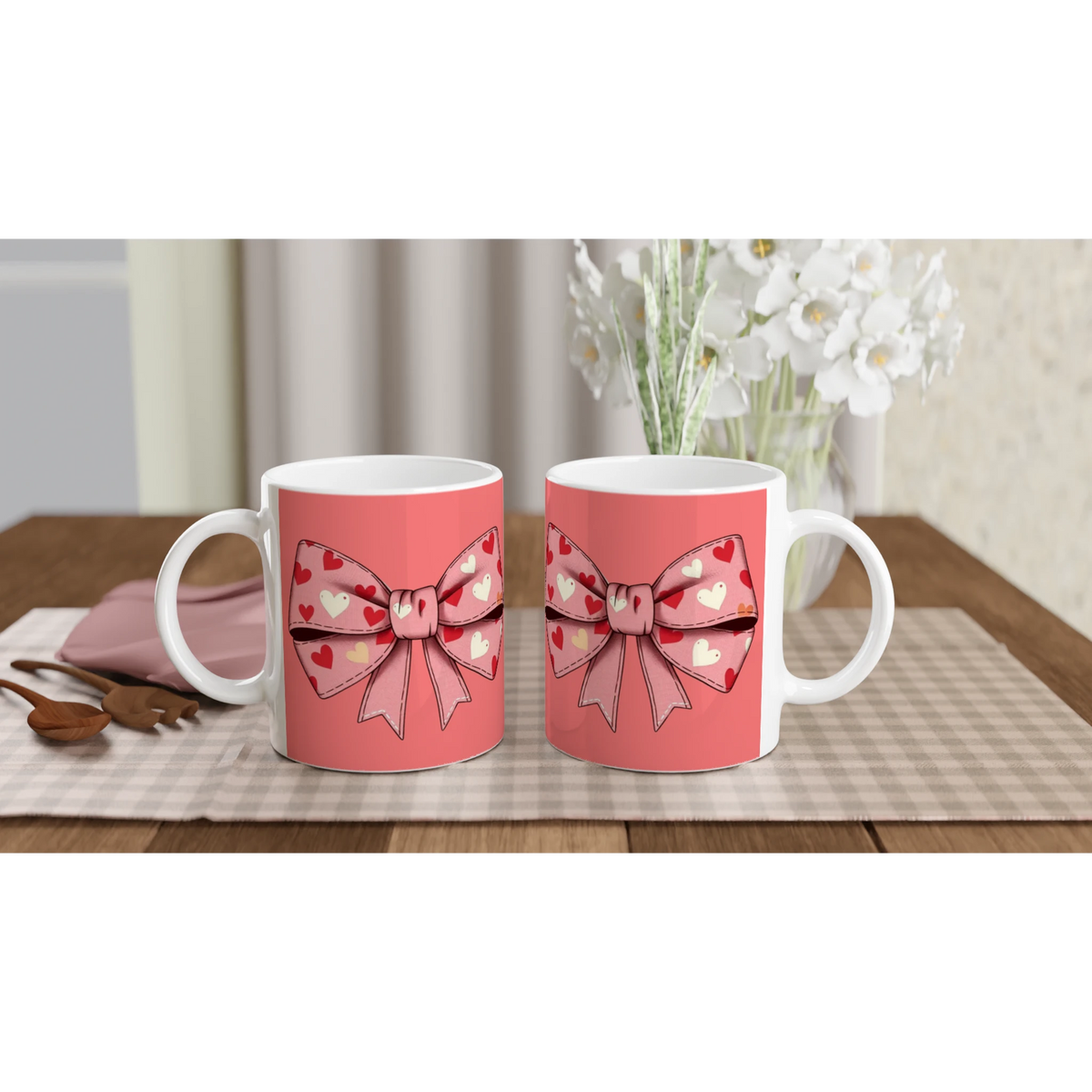 Two pink mugs with a heart-shaped bow design on a table, white flowers in the background.