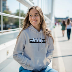 Young woman in a light gray hoodie featuring Believe In Yourself slogan, smiling outdoors.