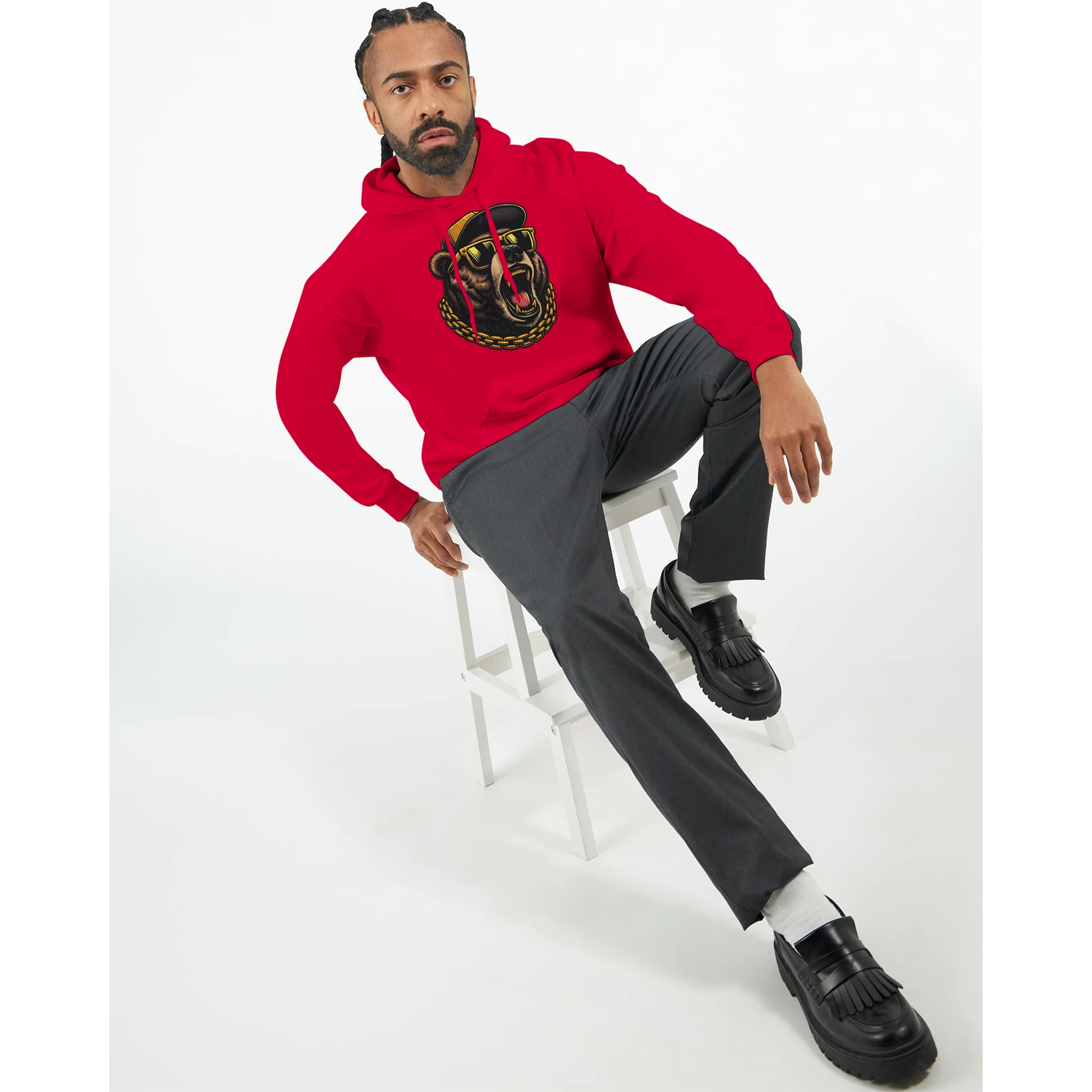 Man wearing red Grizzly Bear Hoodie with graphic design, seated on white stool, grey pants, black loafers.