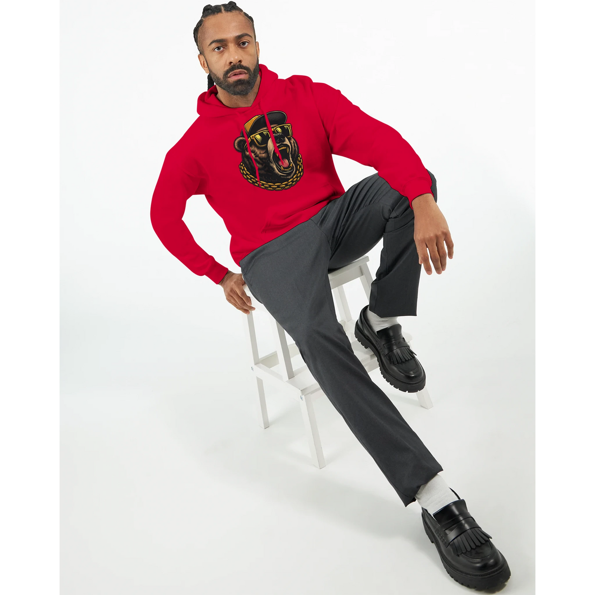 Man wearing red Grizzly Bear Hoodie with graphic design, seated on white stool, grey pants, black loafers.