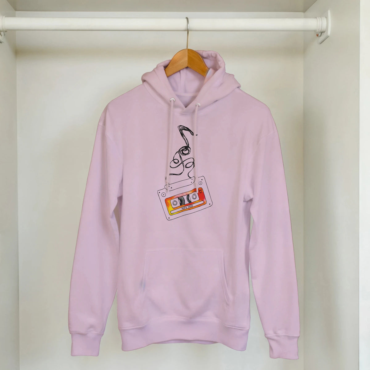 Pink hoodie featuring a retro cassette tape graphic with "90s Hits" label and swirling musical notes, hanging on a wooden hanger.