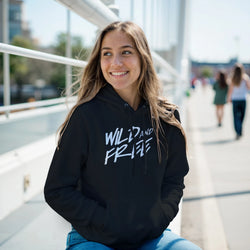 Woman wearing Cute Wild And Free Hoodie - Cozy Slogan Top for Women, black with bold white text on city street.