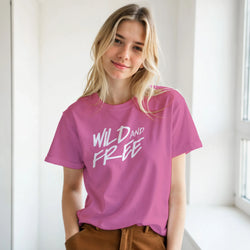 Woman wearing pink 'Wild and Free' slogan T-shirt, retro streetwear vibe by a bright window.