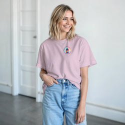 Woman wearing a soft pink retro graphic tee with a small circular palm tree chest logo, styled casually with blue denim jeans.