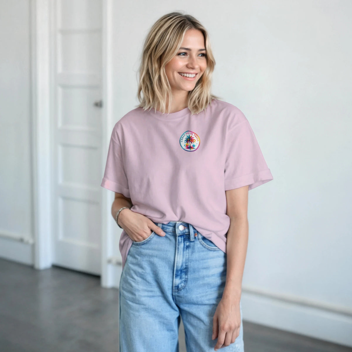 Woman wearing a soft pink retro graphic tee with a small circular palm tree chest logo, styled casually with blue denim jeans.