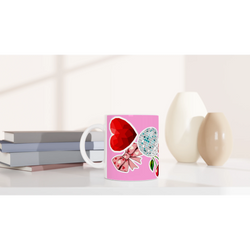 Pink ceramic coffee mug featuring a red heart, disco ball, cherries, and bow design on a white desk with books.