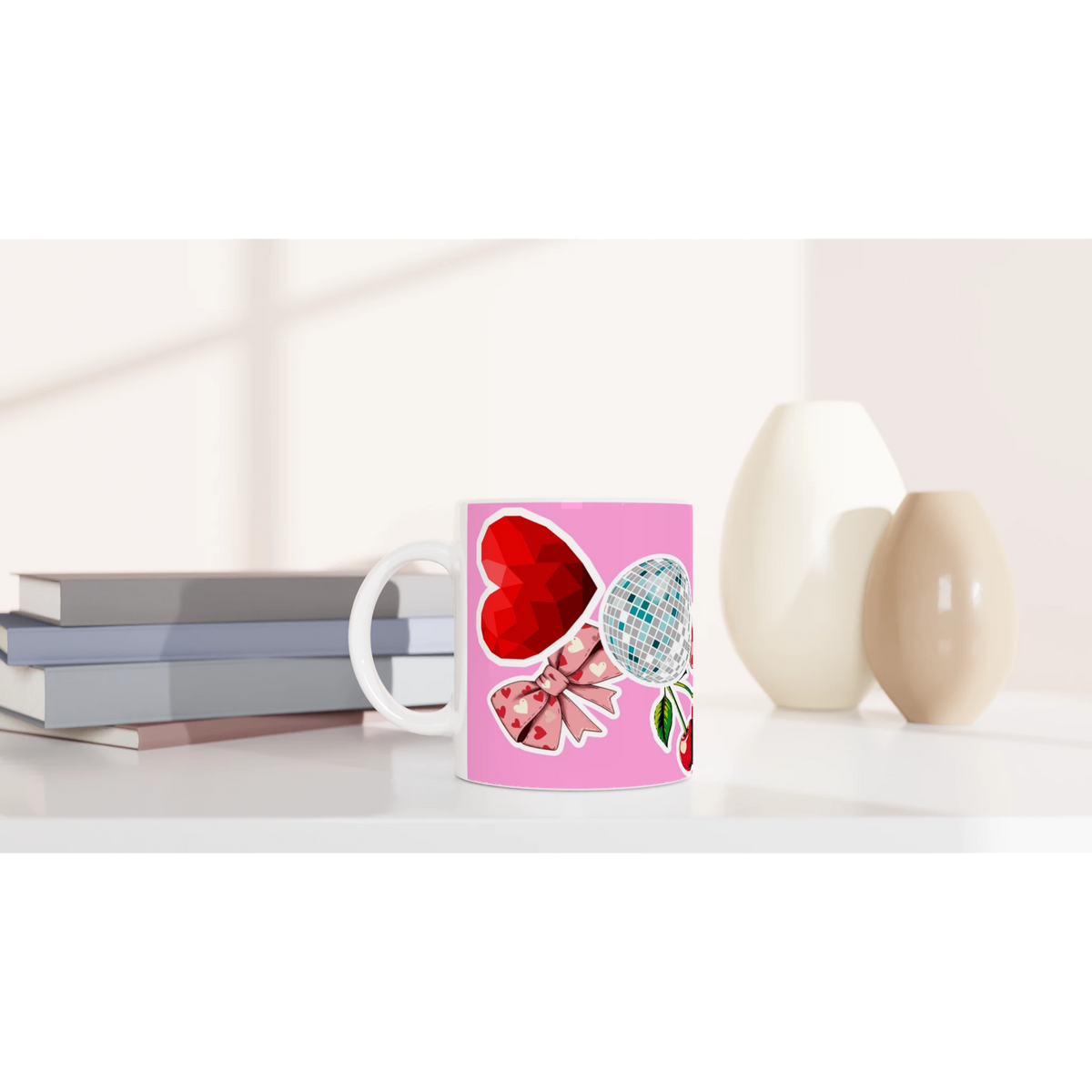 Pink ceramic coffee mug featuring a red heart, disco ball, cherries, and bow design on a white desk with books.