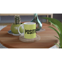 Light-green mug with bold POSITIVE text sits on a wooden table beside a small cactus.