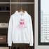 White hoodie with 'Sweet Cherries' design on a hanger in a room with shelves and a window.