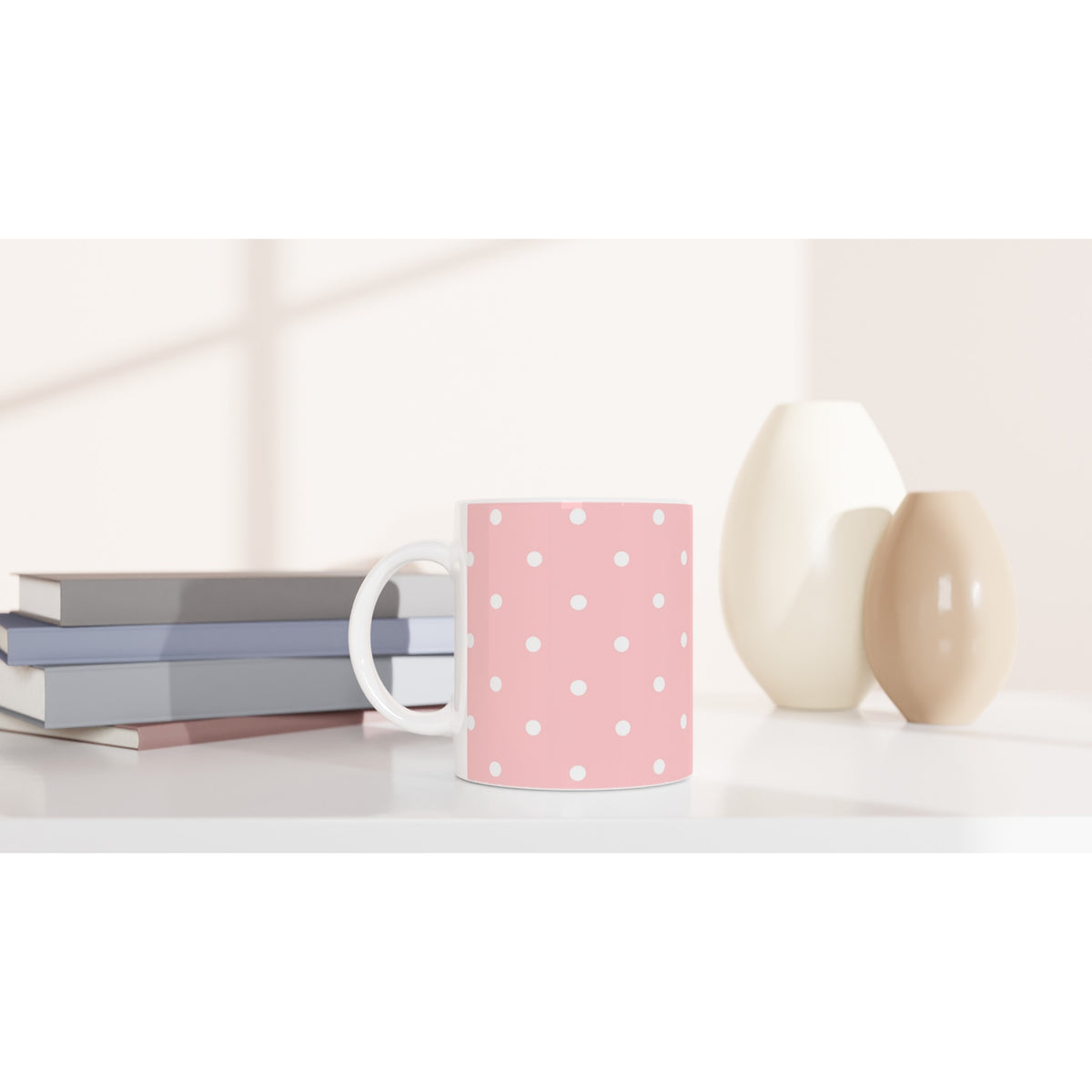 Pastel pink and white polka dot ceramic mug on a white desk beside stacked books and minimalist vases.