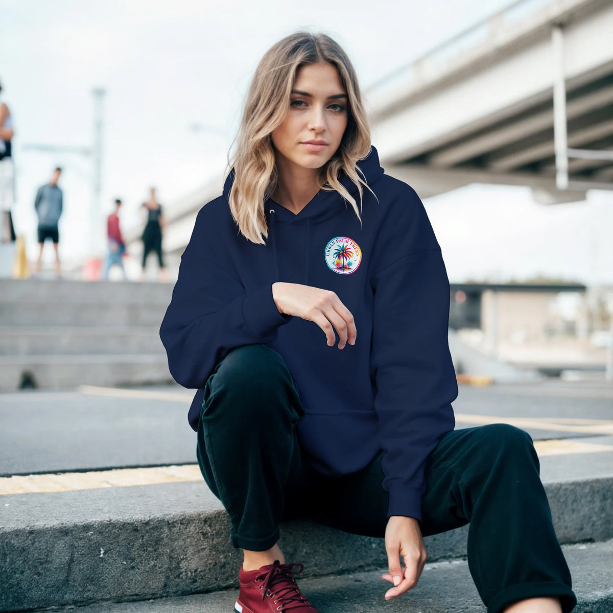 Woman in navy retro palm tree graphic hoodie crouching on urban steps. Casual streetwear look with relaxed fit and colorful chest logo.