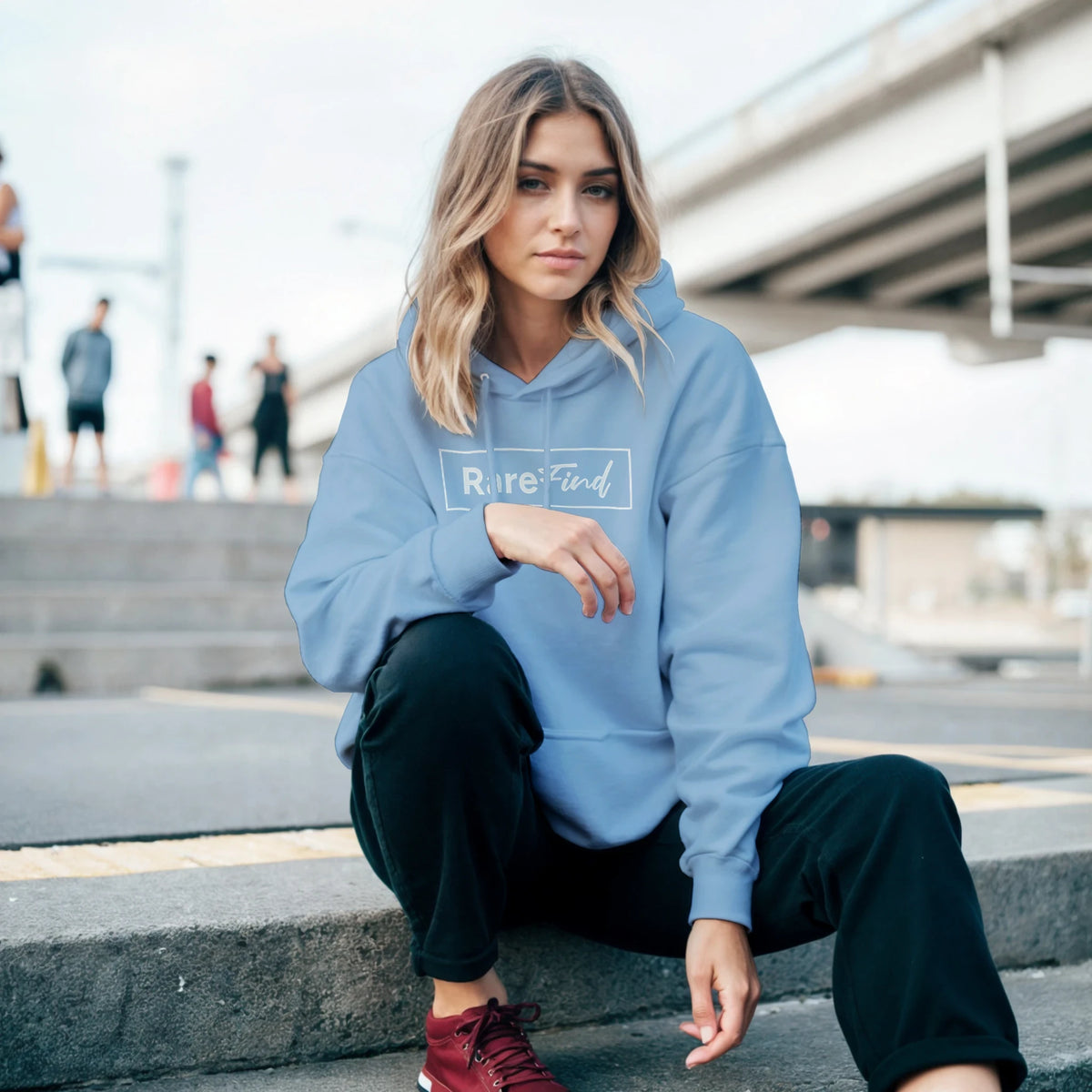 Woman in a light blue hoodie with front pouch pocket and Rare Find slogan, seated on outdoor steps.