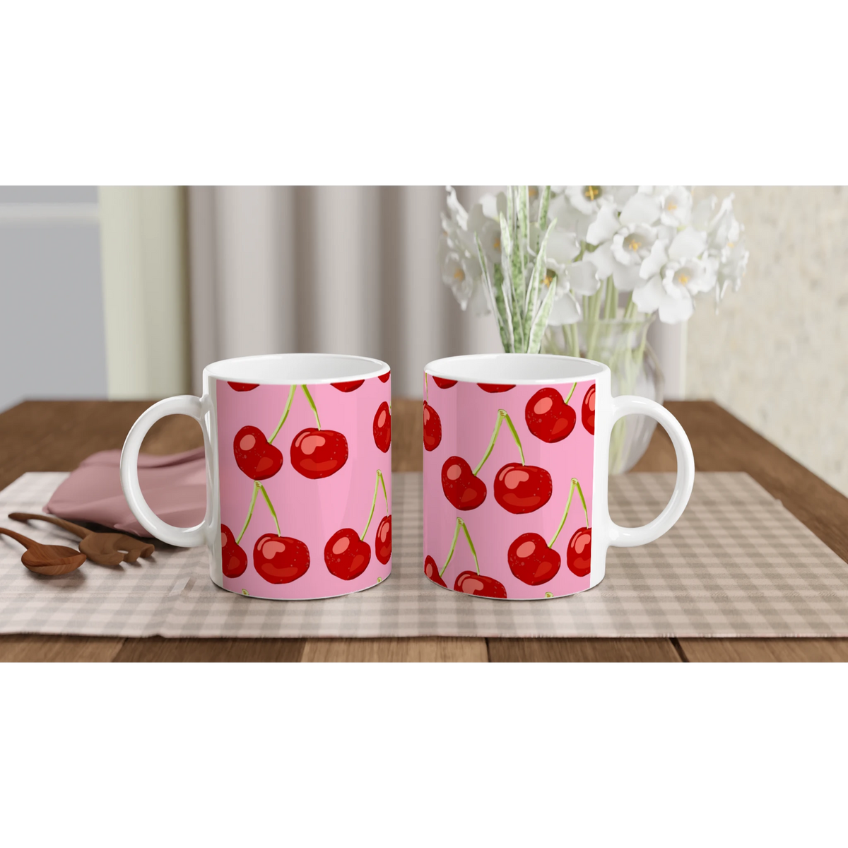 Two pink cherry-pattern ceramic mugs with white handles on a checkered table, cozy kitchen scene.
