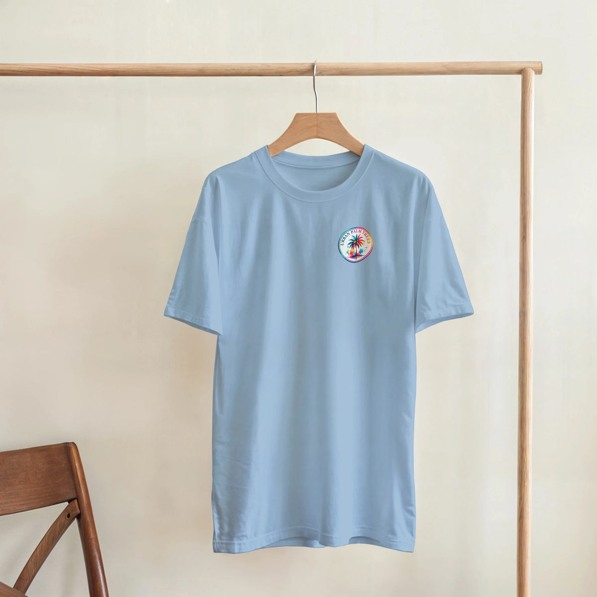 Light blue t-shirt with small retro palm tree graphic on chest, hanging on wooden rack.