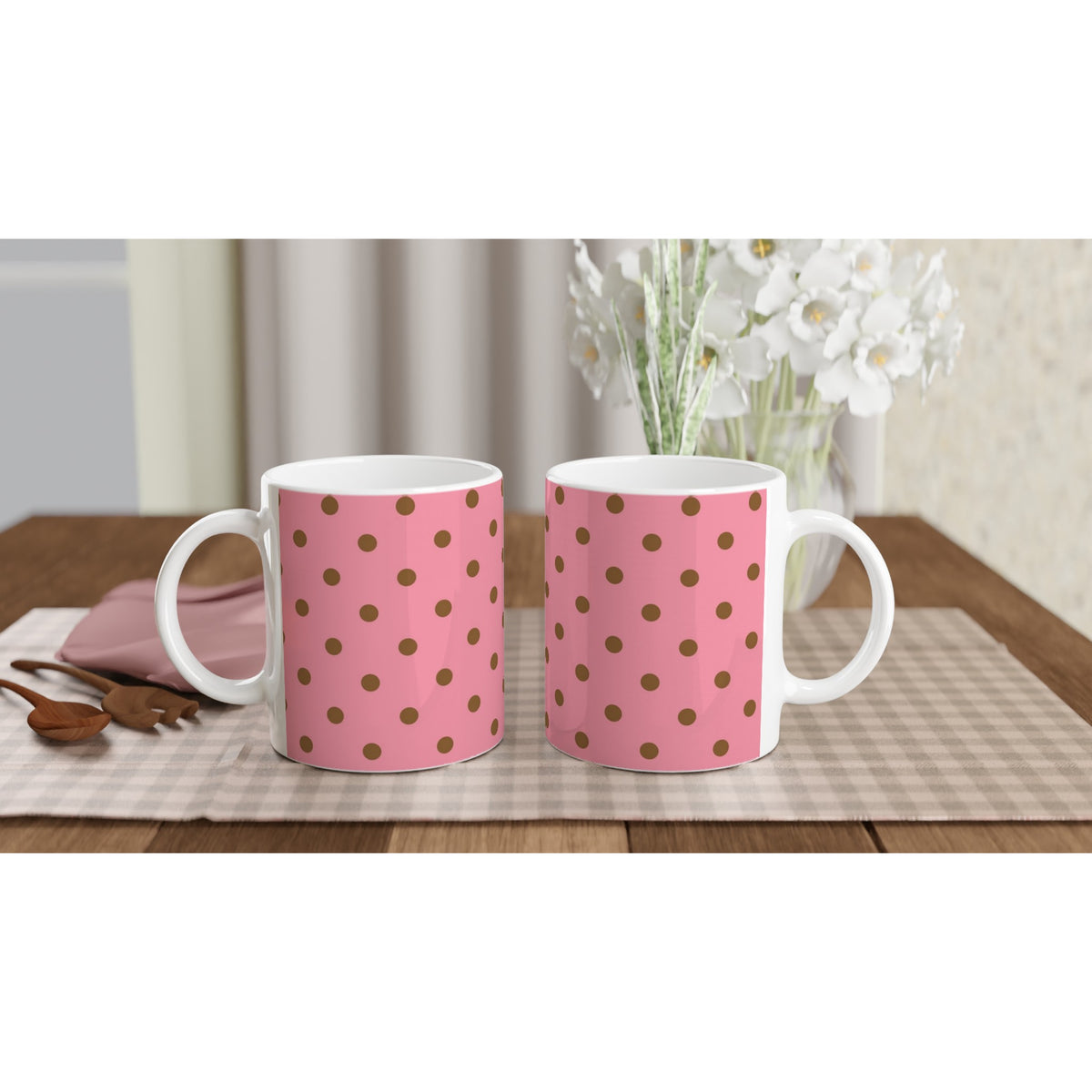 Two pink ceramic mugs with brown polka dots sit on a checkered mat next to wooden spoons and white flowers.