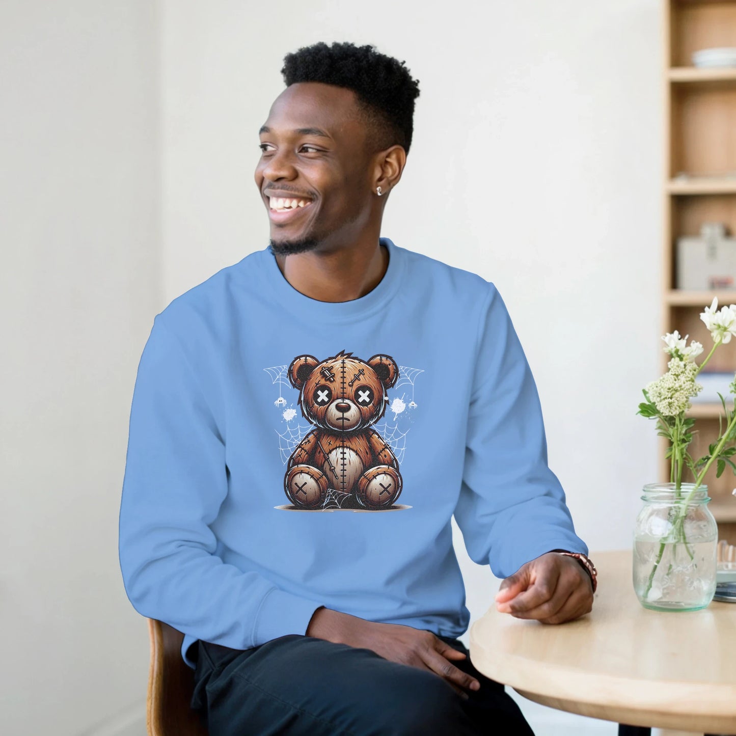 Person wearing a blue men's graphic sweatshirt with a teddy bear graphic, sitting in a room with a table and flowers.