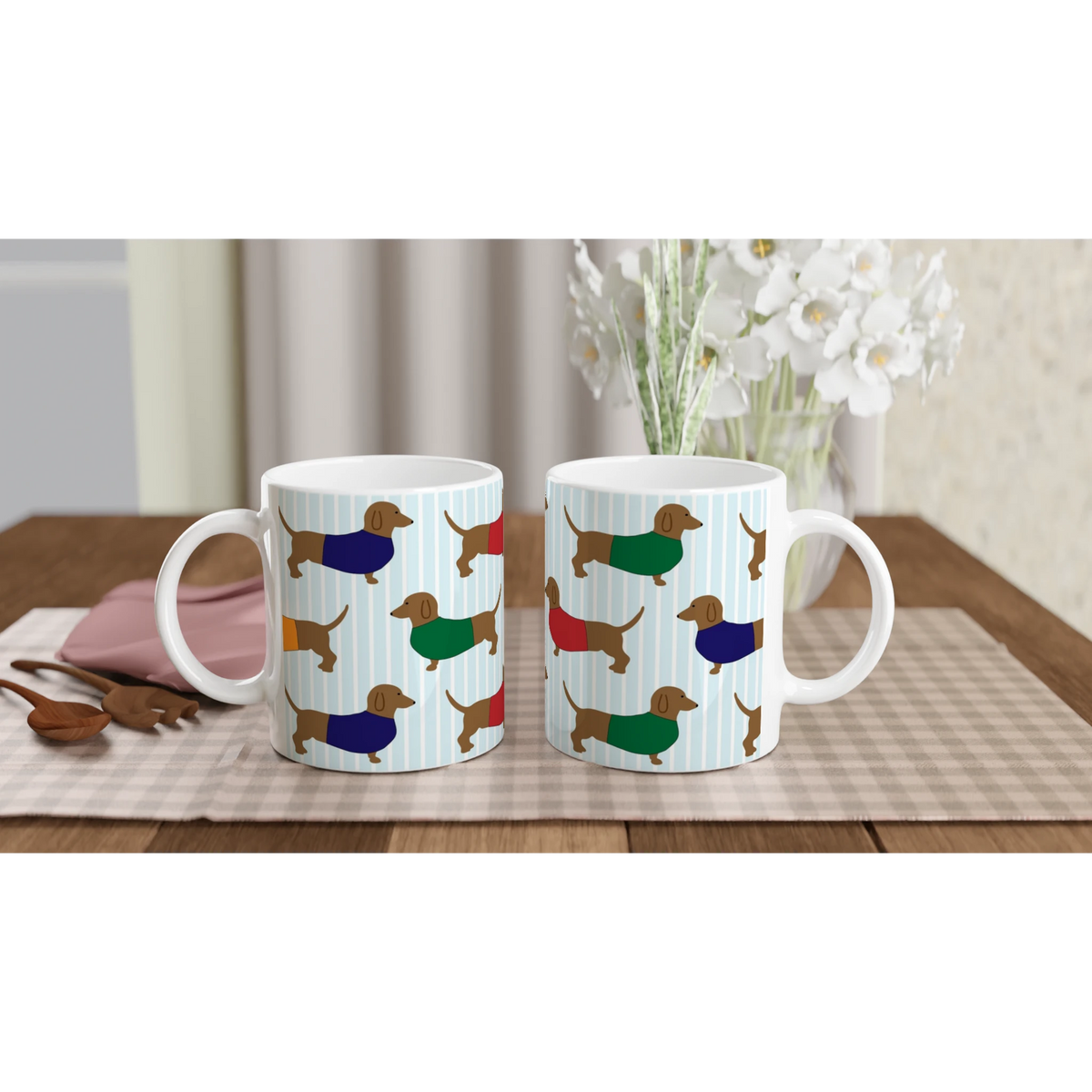 Two dachshund-pattern ceramic mugs with blue stripes, set on a table with a vase of white flowers.