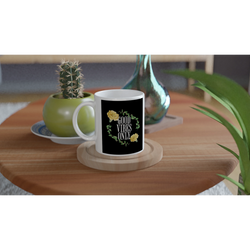 White ceramic mug with black label reading GOOD VIBES ONLY, surrounded by a green leafy wreath and yellow flowers.