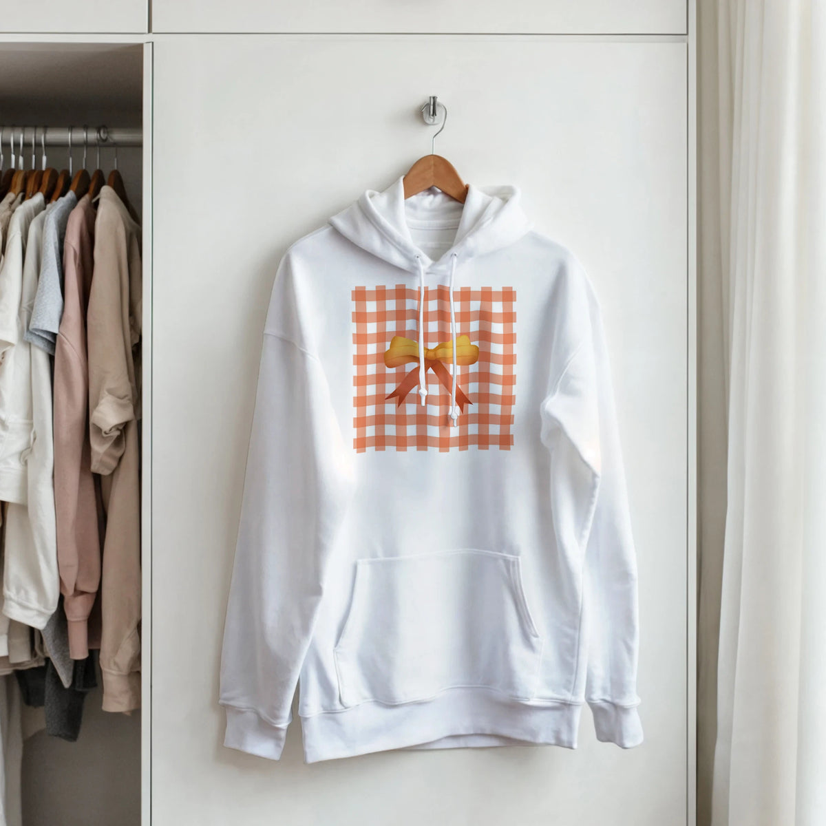 White pullover hoodie featuring an orange gingham square design with a yellow coquette bow, hanging on a closet door.