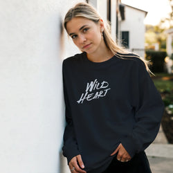 Model wearing Cute Wild Heart sweatshirt with white script slogan beside a white wall outdoors.