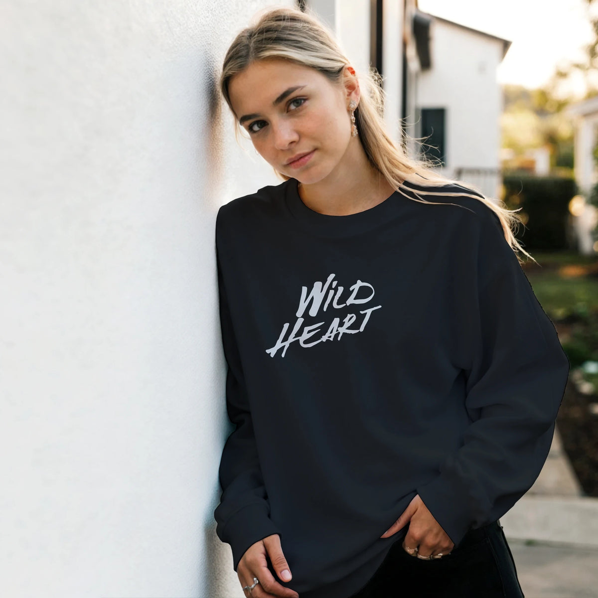 Model wearing Cute Wild Heart sweatshirt with white script slogan beside a white wall outdoors.