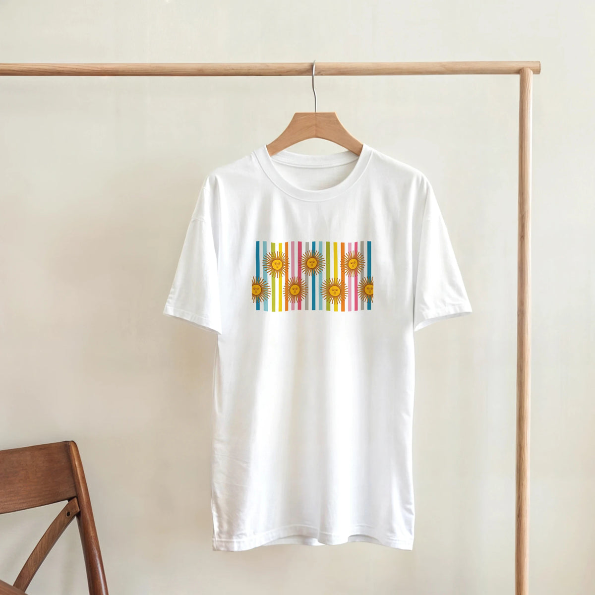 White women's T-shirt on a wooden hanger featuring a retro rainbow sun graphic with vertical color bars.