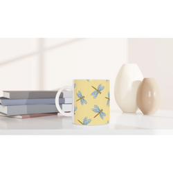 Yellow dragonfly mug with blue dragonfly illustrations sits on a desk beside stacked books.