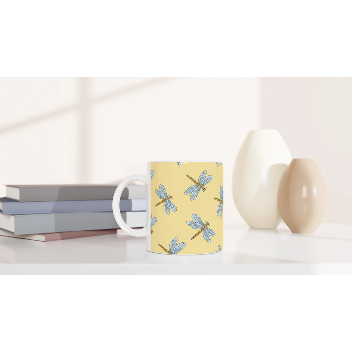 Yellow dragonfly mug with blue dragonfly illustrations sits on a desk beside stacked books.