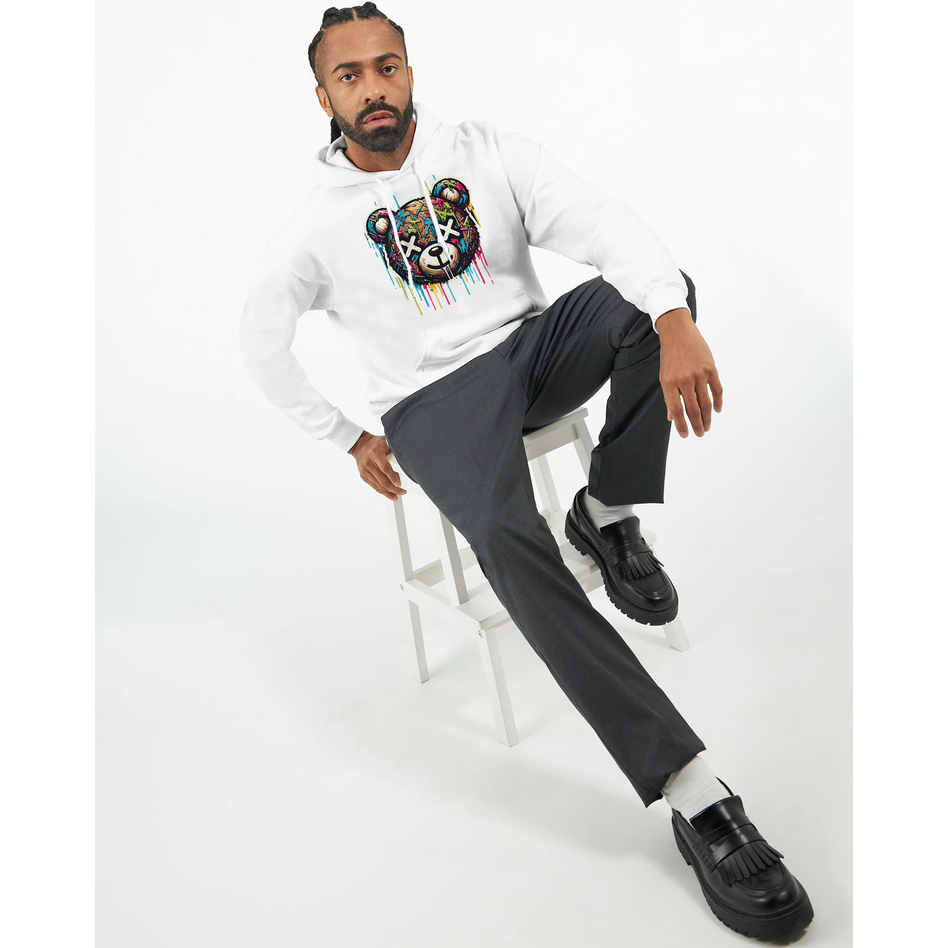 Man sitting on a stool wearing a white men's teddy bear hoodie with a colorful design, gray pants, and black shoes.