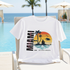 White t-shirt with a tropical graphic design on a lounge chair by a pool with palm trees and blue sky.